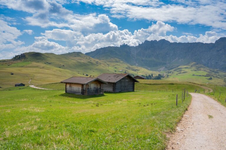 Which hotels on the Seiser Alm are best for a hiking holiday with trails starting right at the door? A wooden alpine hut beside a gravel hiking path across green meadows with mountains behind